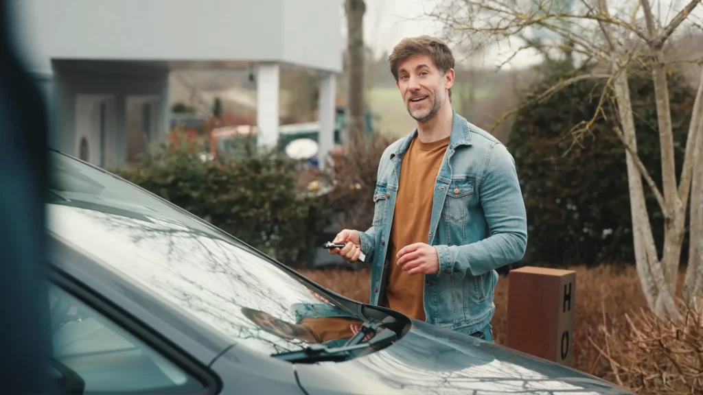 Mann in Jeansjacke spricht mit Autoschlüssel in der Hand vor einem Auto in die Kamera, Szene aus einem Werbefilm-Dreh